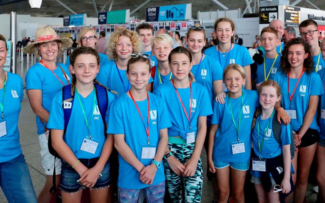 A large group of around Cambridge Dive Team divers assembled at an airport. All the divers are wearing light blue CDT tee-shirts and the majority are children.