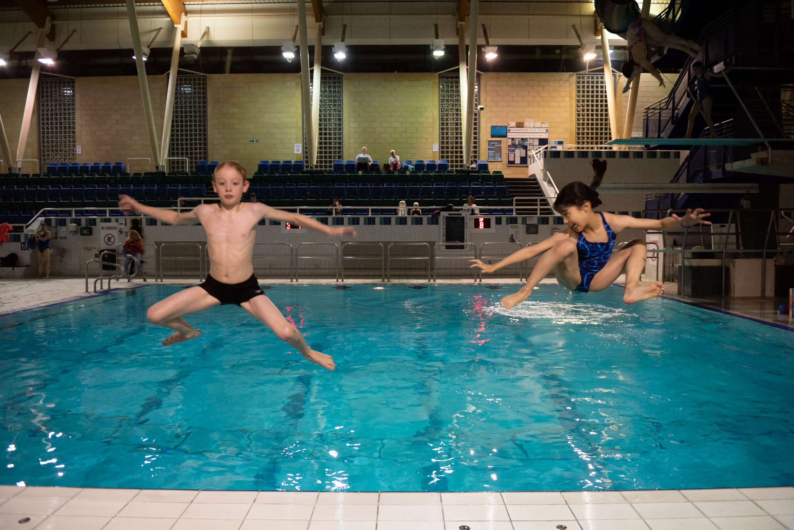 A boy and a girl enjoying frivolously jumping into a diving pool from poolside. Each child is in a star position.