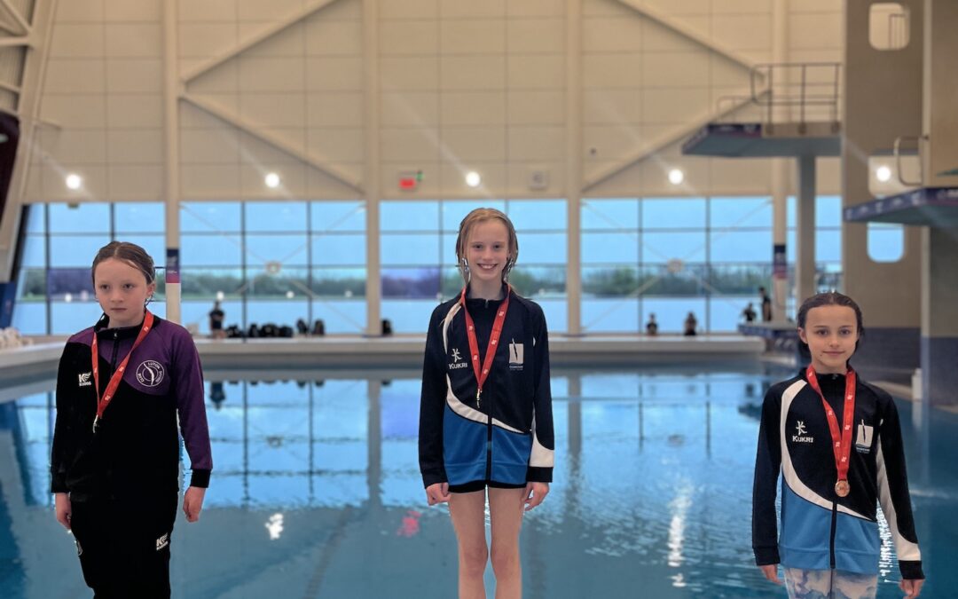 Three young divers standing on a podium on poolside, wearing medals.